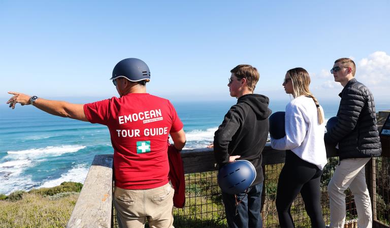 A tour guide wearing a branded red Tshirt and helmet points to the distance while a tour group of three adults look on.