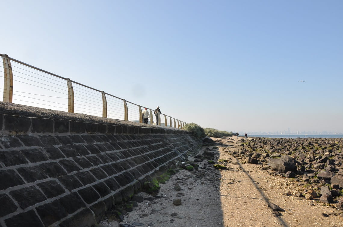 The repaired bluestone seawall at Point Gellibrand Coastal Heritage park runs along the left of the image, with two people walking along the top of it, fenced in by balustrade. To the right of the image is a rocky beach, and the water and city skyline is in the distance. 