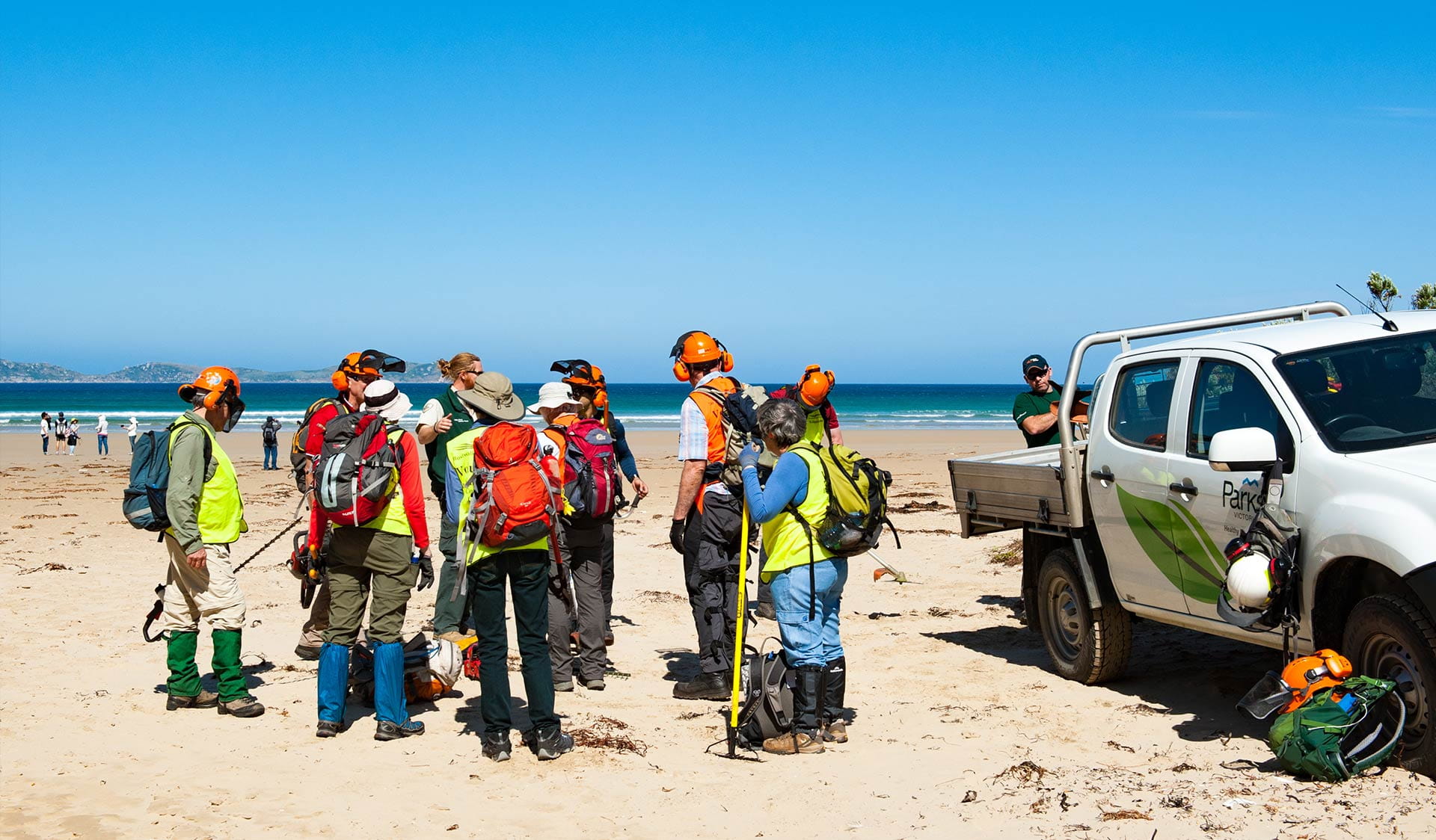 Group of people in high visibility vests gathering next to a Parks Victoria vehicle on a beach